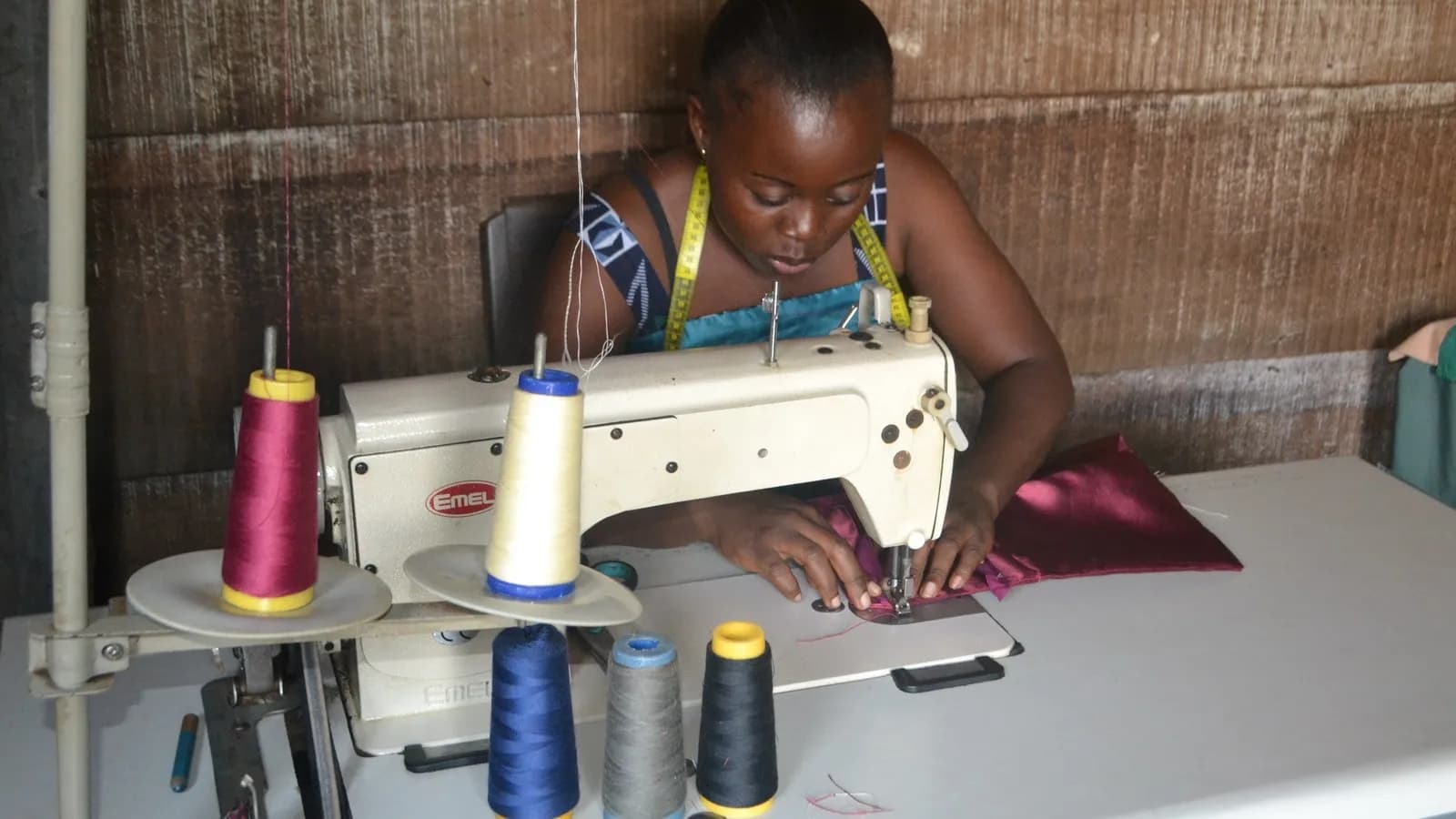 A seamstress working at an industrial sewing machine in her workshop, cones of thread beside her and a tape measure round her neck