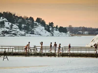 Winter swimmers in swimsuits on a wooden pier beside a hole cut in the sea ice at Turku, Finland, in low afternoon sun