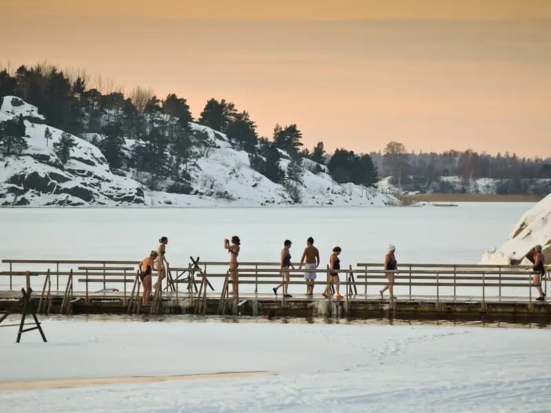 Winter swimmers in swimsuits on a wooden pier beside a hole cut in the sea ice at Turku, Finland, in low afternoon sun