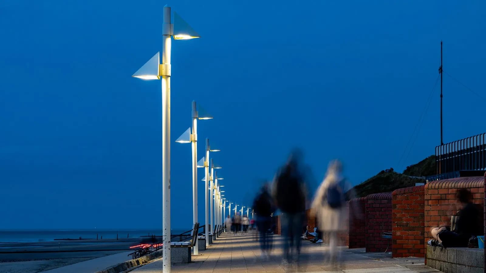 People walking along a lit seafront promenade at dusk, slightly blurred by the long exposure, with the beach to the left