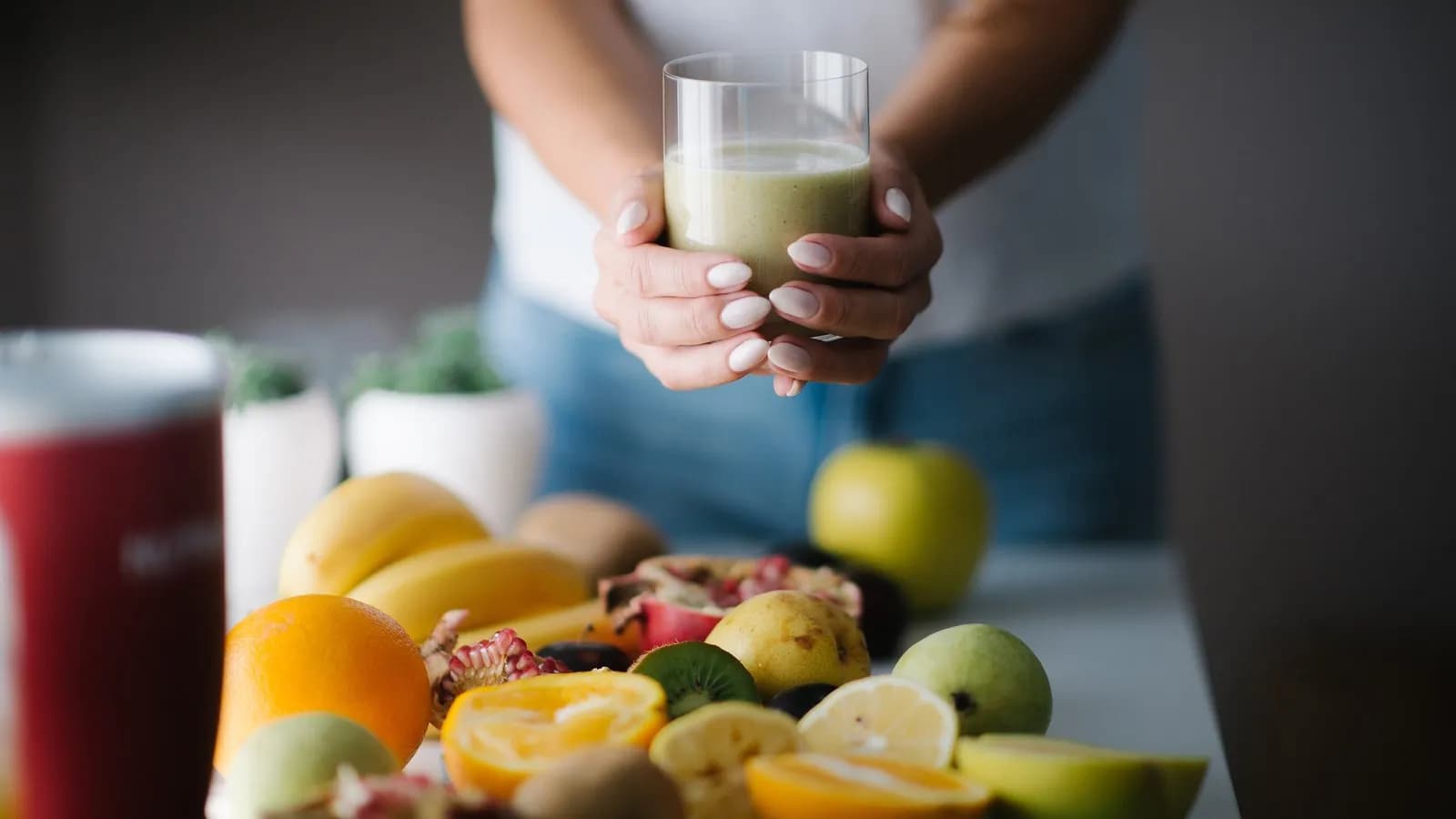 A person holding a glass of green smoothie behind a table of cut fruit