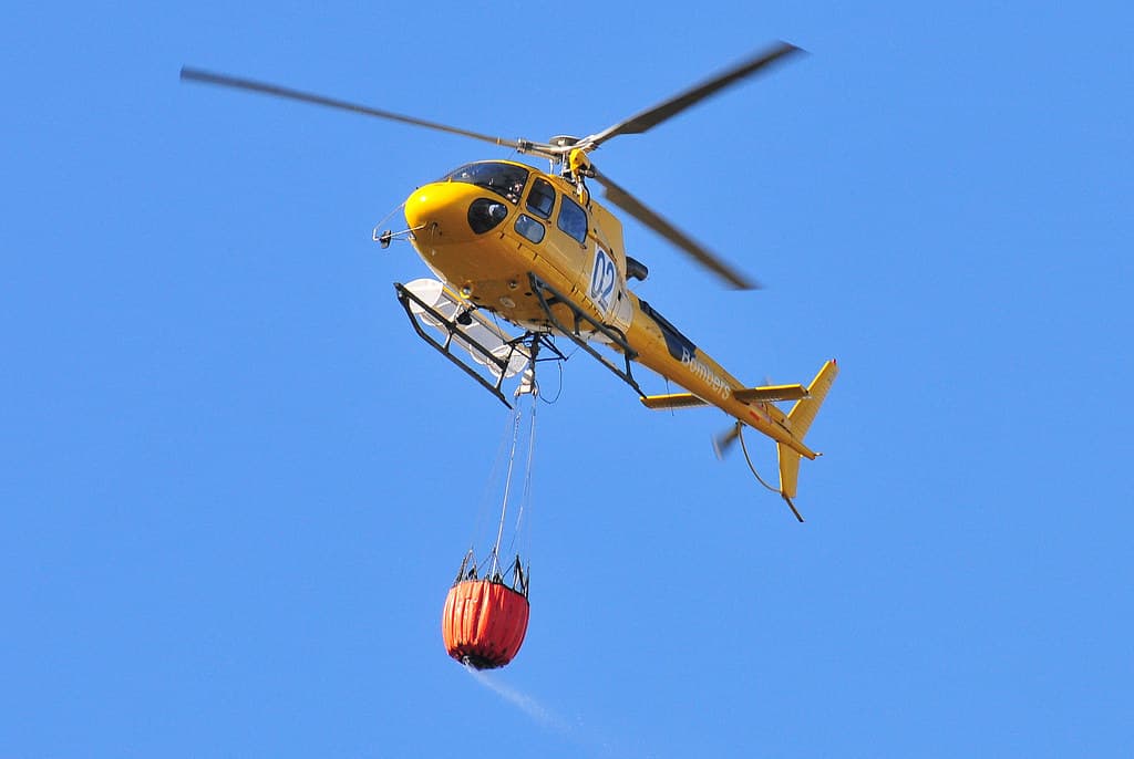 A firefighting helicopter dropping water near a smoke-covered European forest and town