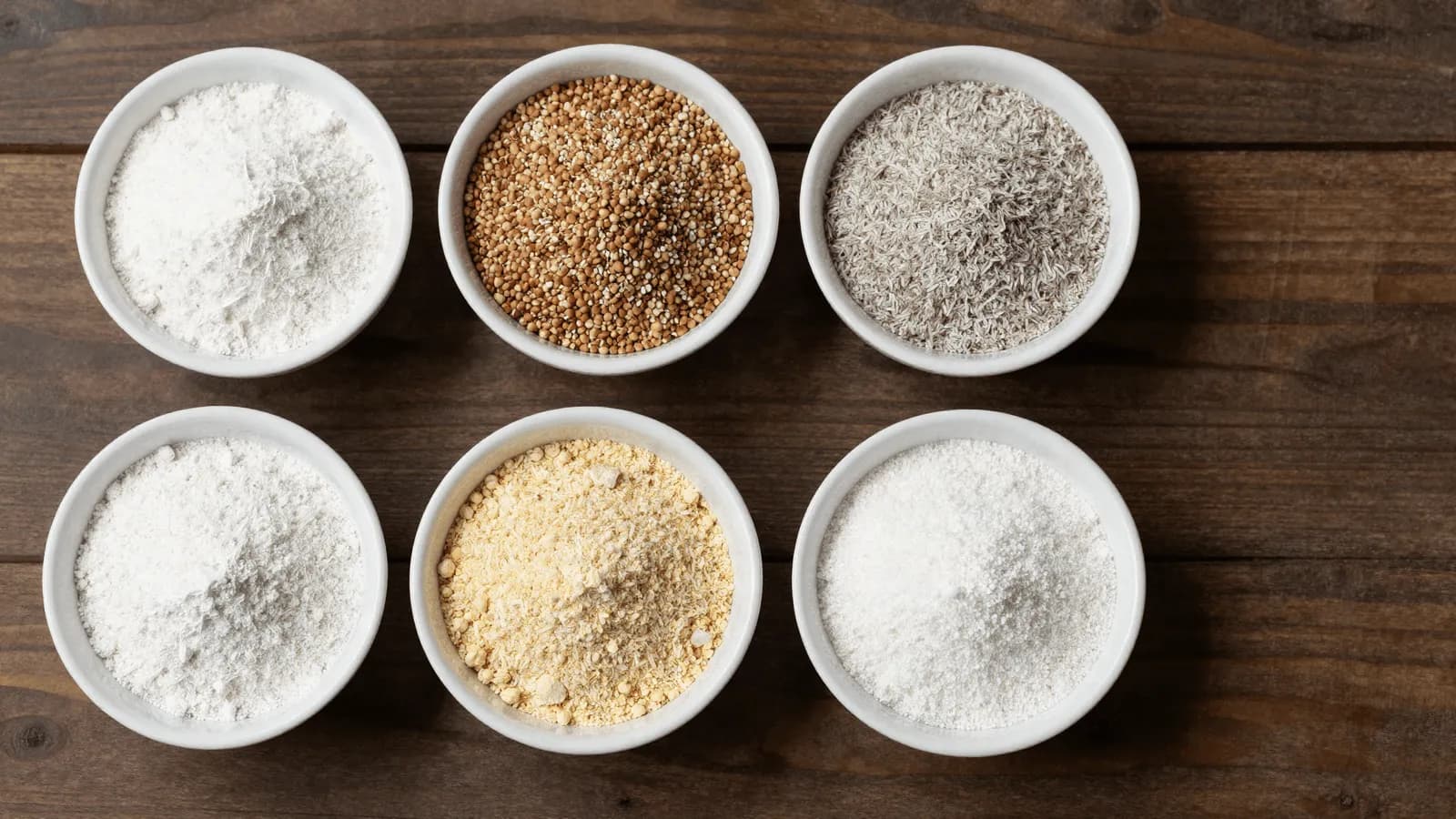 Small bowls of different gluten-free flours arranged on a wooden surface