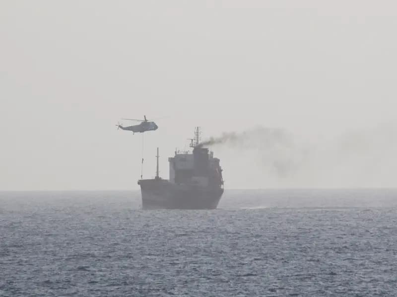 A merchant tanker under way in the Arabian Gulf while a helicopter lowers a boarding party onto its deck. The photograph records the boarding of the MT Wila in August 2020 and is not from the current closure