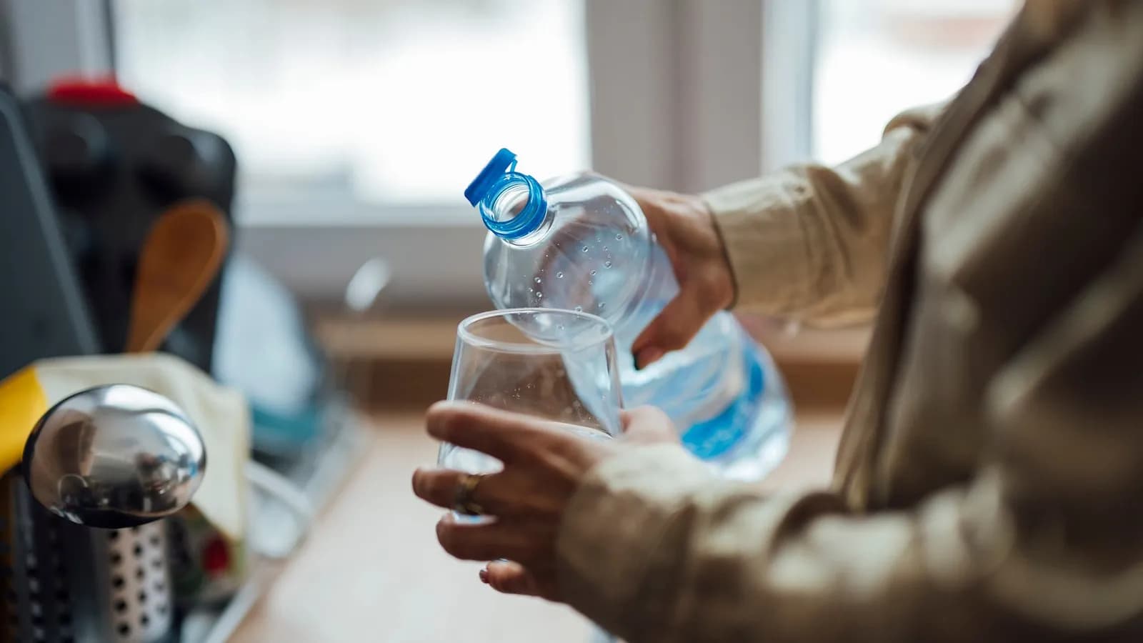 Water being poured from a bottle into a drinking glass held in the hand, at a kitchen worktop by a window