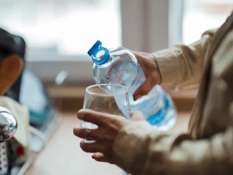 Water being poured from a bottle into a drinking glass held in the hand, at a kitchen worktop by a window