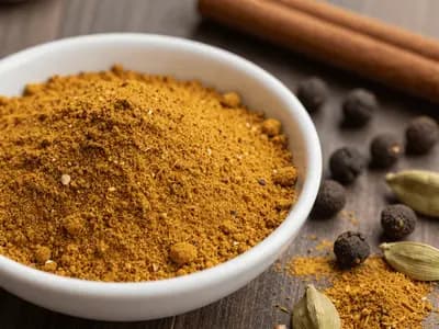 A small bowl of warm brown-gold ground spice blend beside whole allspice berries and a cinnamon stick