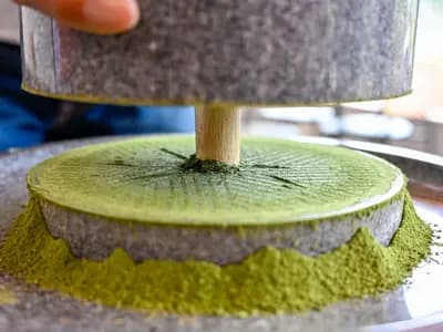 A pale jade green matcha latte being poured into a white ceramic cup on a cafe counter beside a bamboo whisk and tin of matcha powder