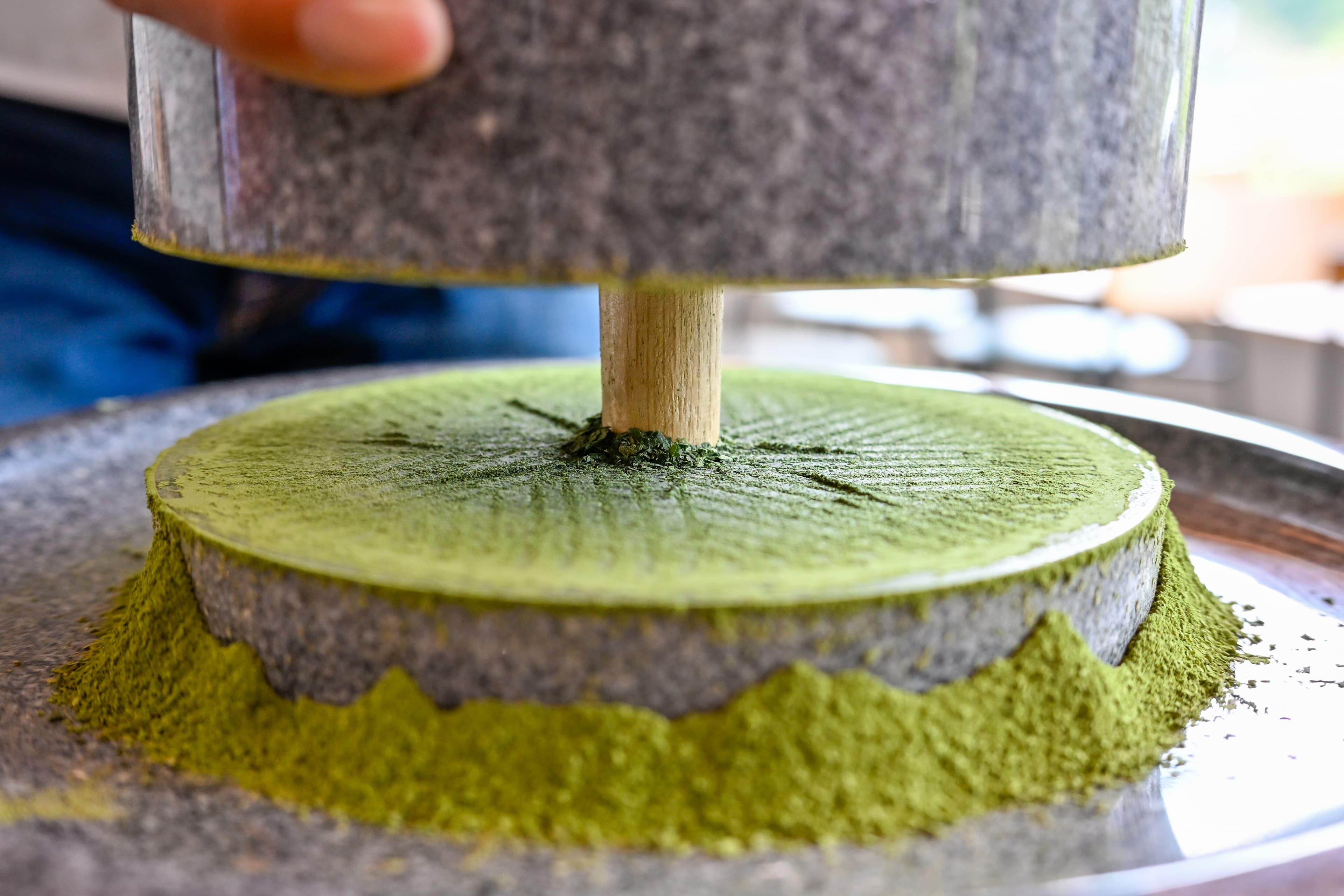 A pale jade green matcha latte being poured into a white ceramic cup on a cafe counter beside a bamboo whisk and tin of matcha powder