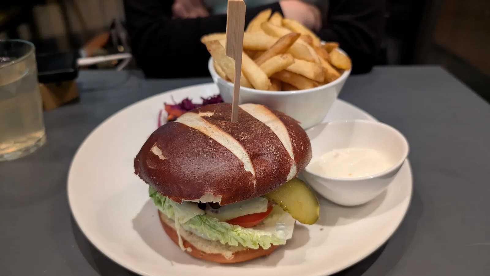 A plant based cheeseburger in a pretzel bun, served with fries, pickles and a pot of mayonnaise