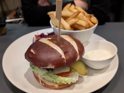 A plant based cheeseburger in a pretzel bun, served with fries, pickles and a pot of mayonnaise