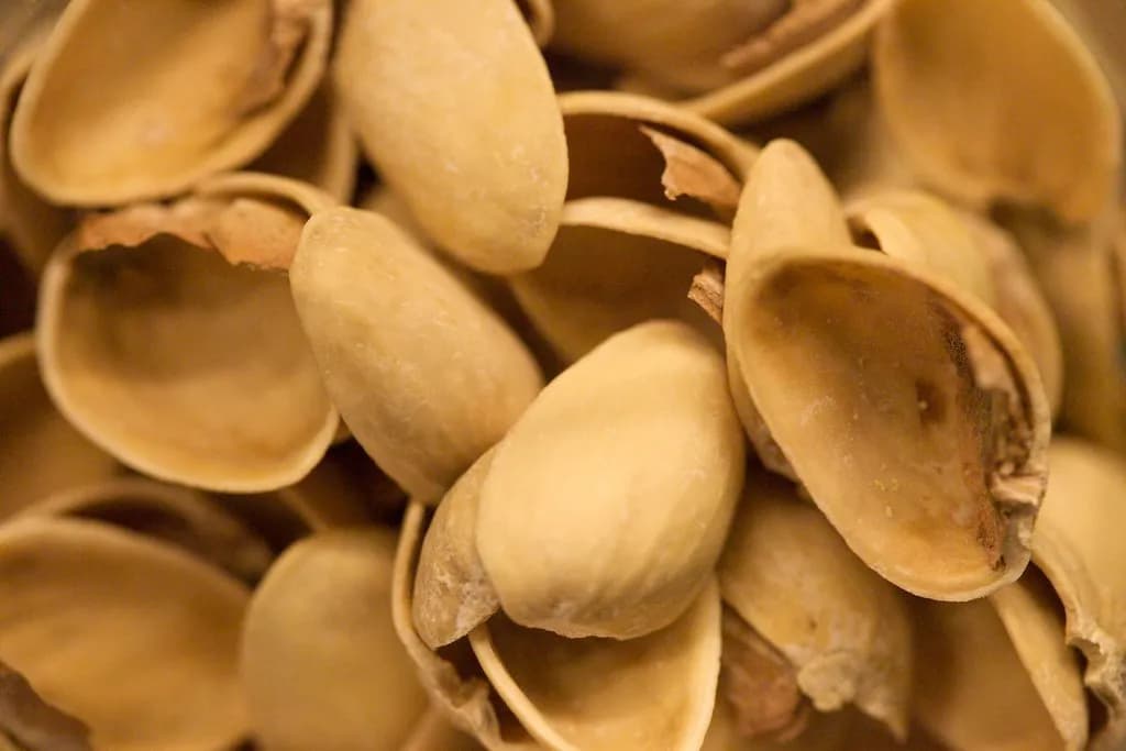 A pile of shelled green pistachios beside a jar of thick pistachio cream paste with a spoon resting in it