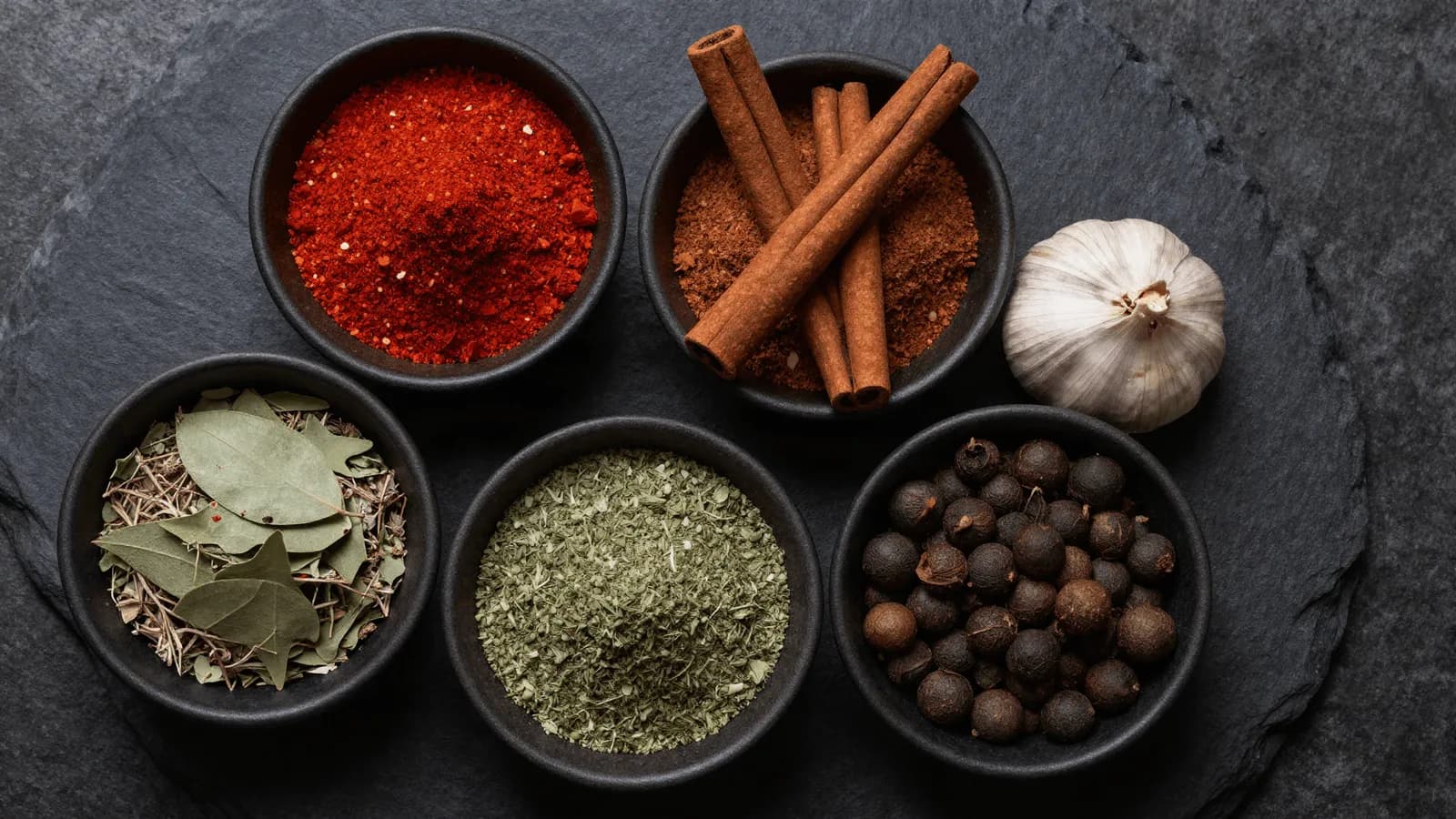 Small bowls of paprika, cinnamon, allspice and dried oregano on a dark surface