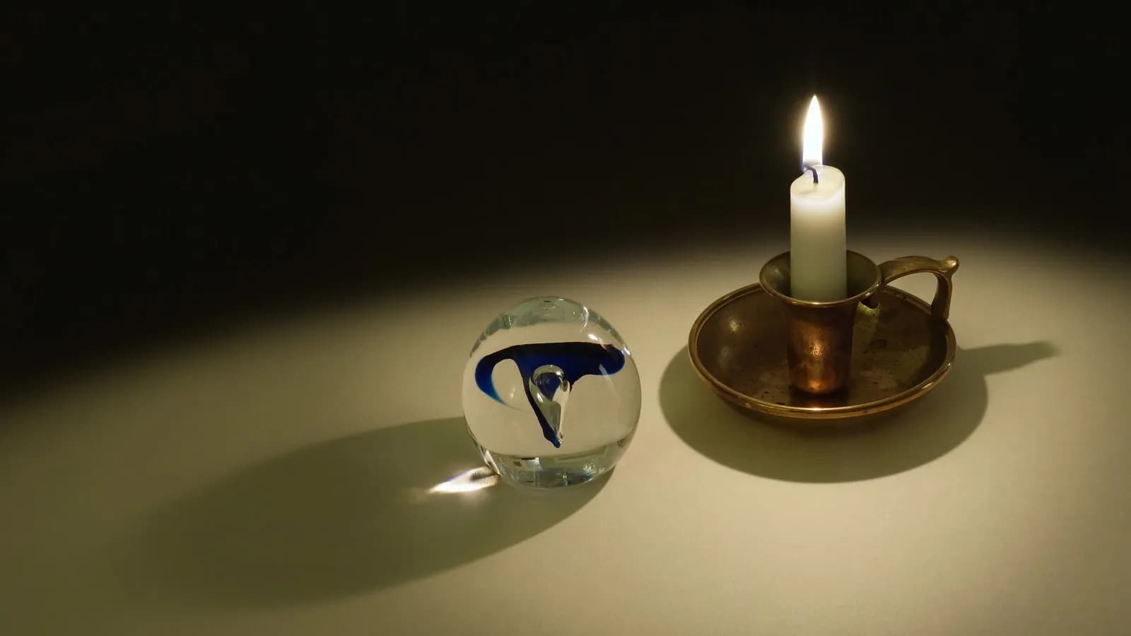 A lit candle in a brass holder on a dark table, its flame refracted through a glass orb beside it