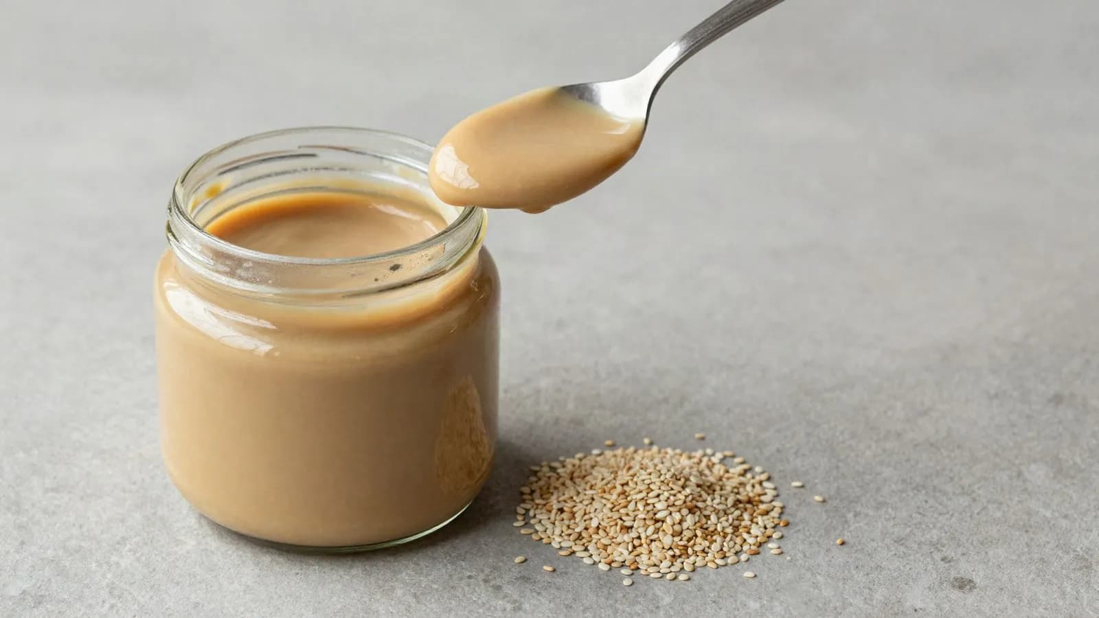 A jar of pale tahini with a spoon lifting a ribbon of the paste