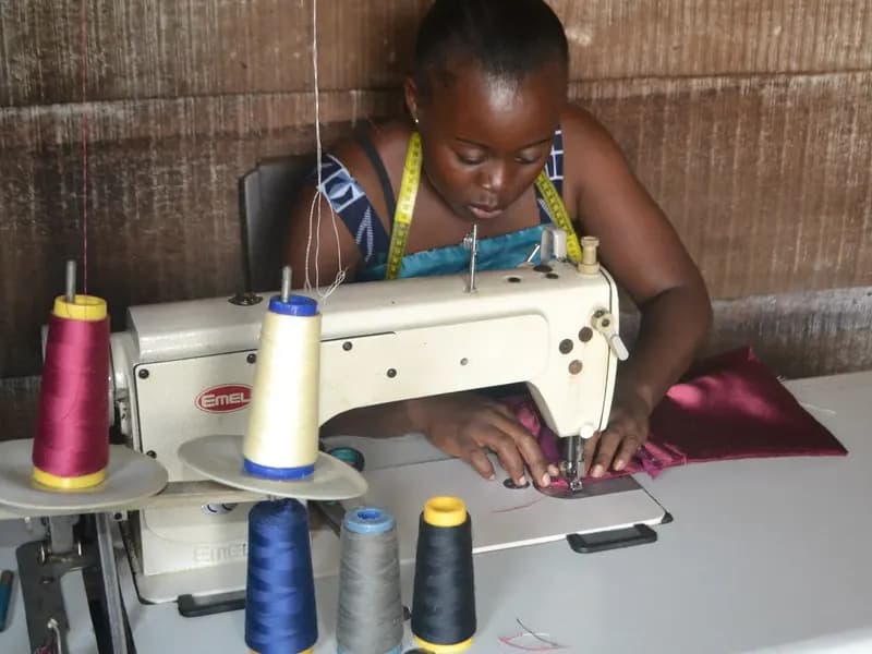 A seamstress working at an industrial sewing machine in her workshop, cones of thread beside her and a tape measure round her neck