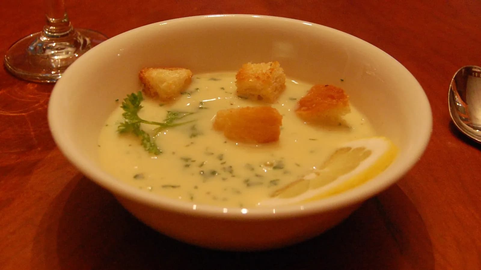 A bowl of pale yellow avgolemono soup with croutons, a sprig of parsley and a slice of lemon