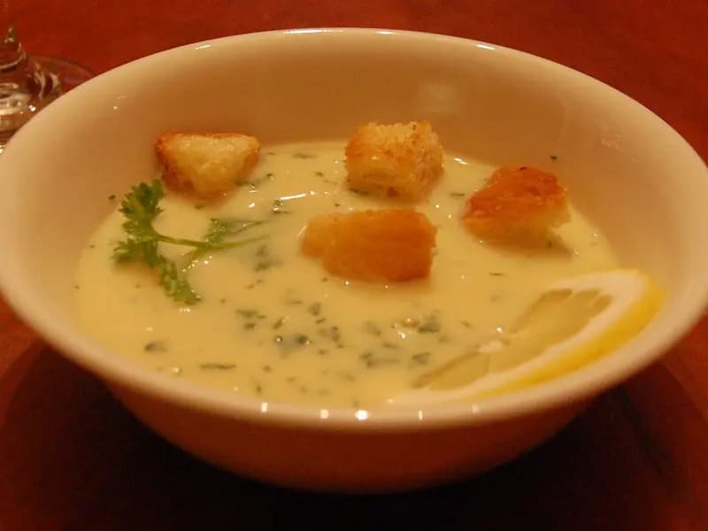 A bowl of pale yellow avgolemono soup with croutons, a sprig of parsley and a slice of lemon