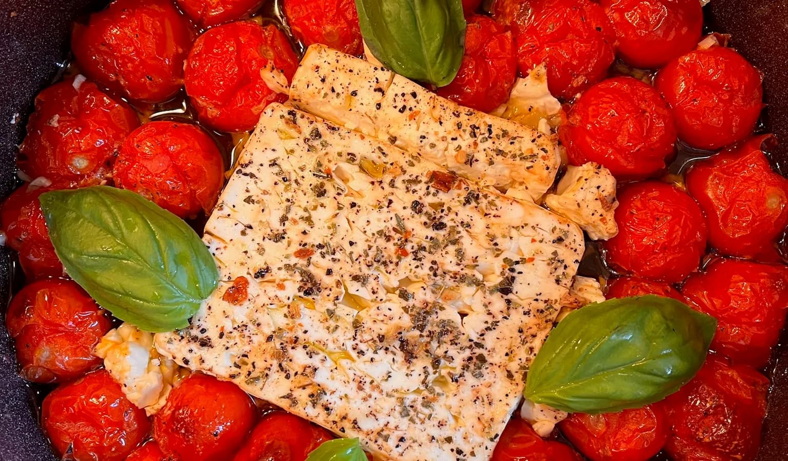 A baking dish with a softened block of feta surrounded by blistered cherry tomatoes, being stirred into a creamy sauce