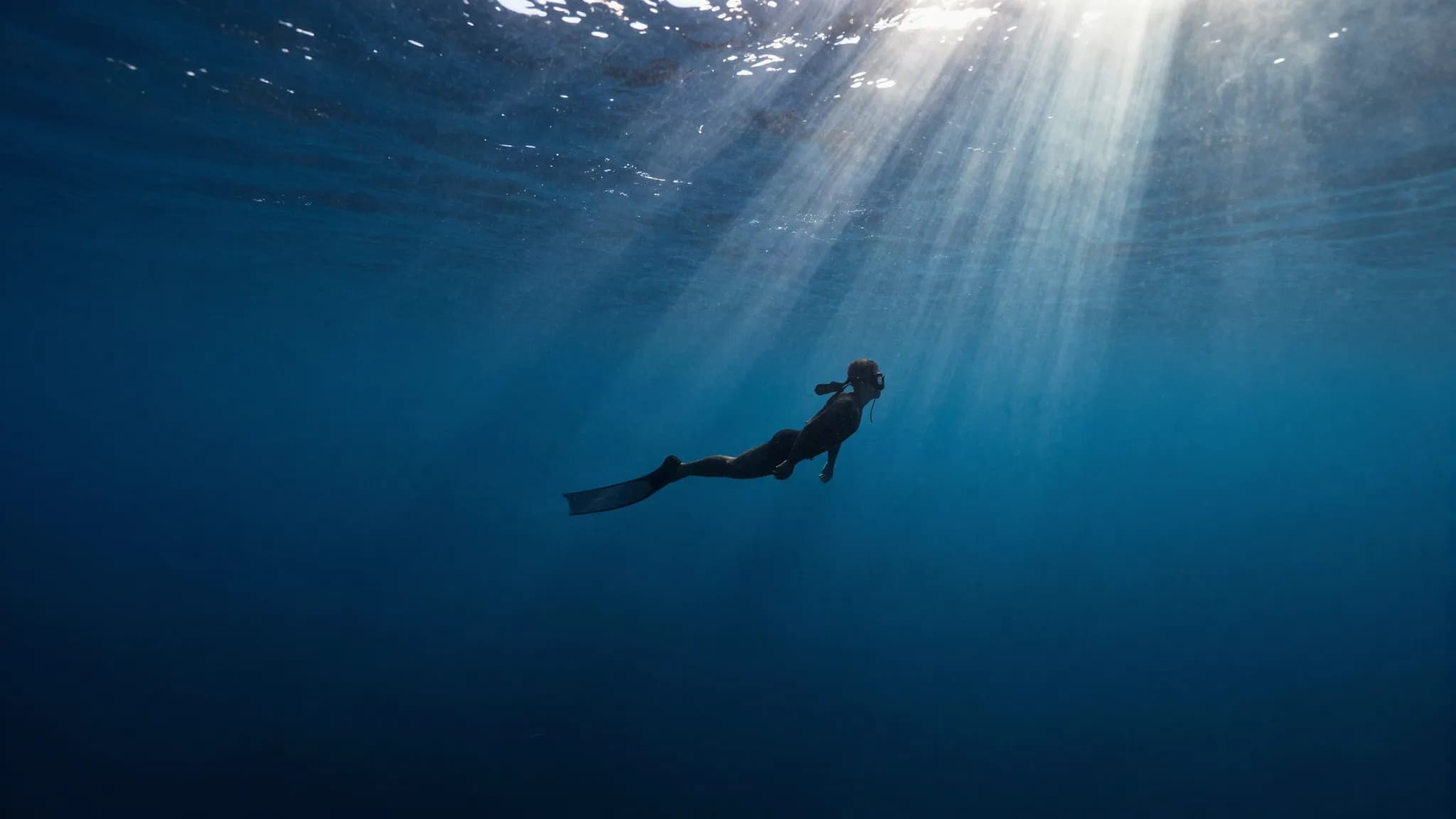 A freediver silhouetted in deep blue water under converging light rays