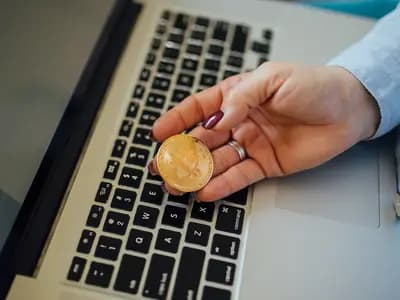 A physical bitcoin token held between finger and thumb in front of an open laptop