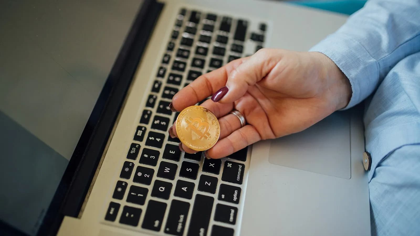A hand holding a gold coloured metal token stamped with the bitcoin logo above a laptop keyboard. Bitcoin has no physical form and tokens like this one are souvenirs rather than currency