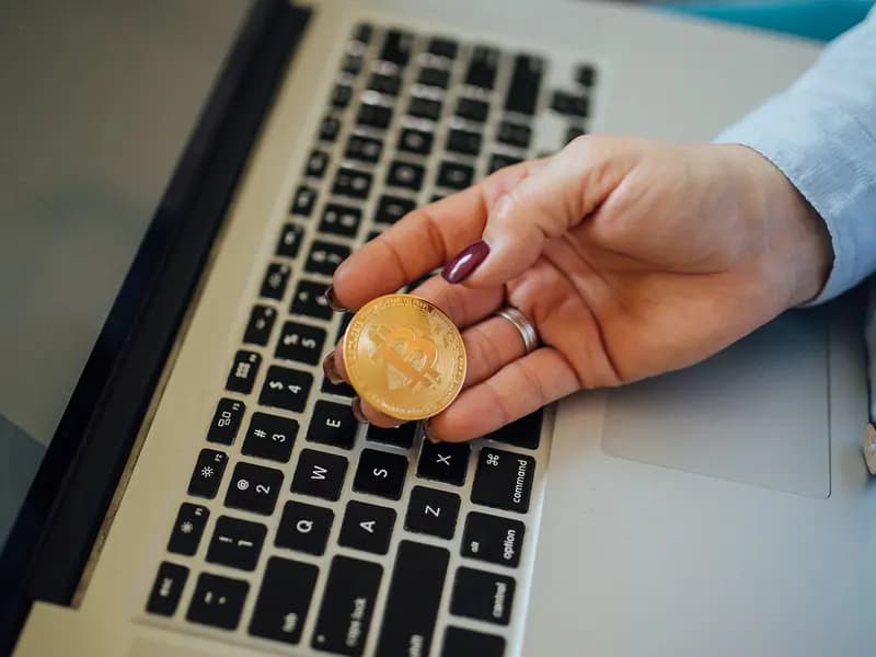 A hand holding a gold coloured metal token stamped with the bitcoin logo above a laptop keyboard. Bitcoin has no physical form and tokens like this one are souvenirs rather than currency