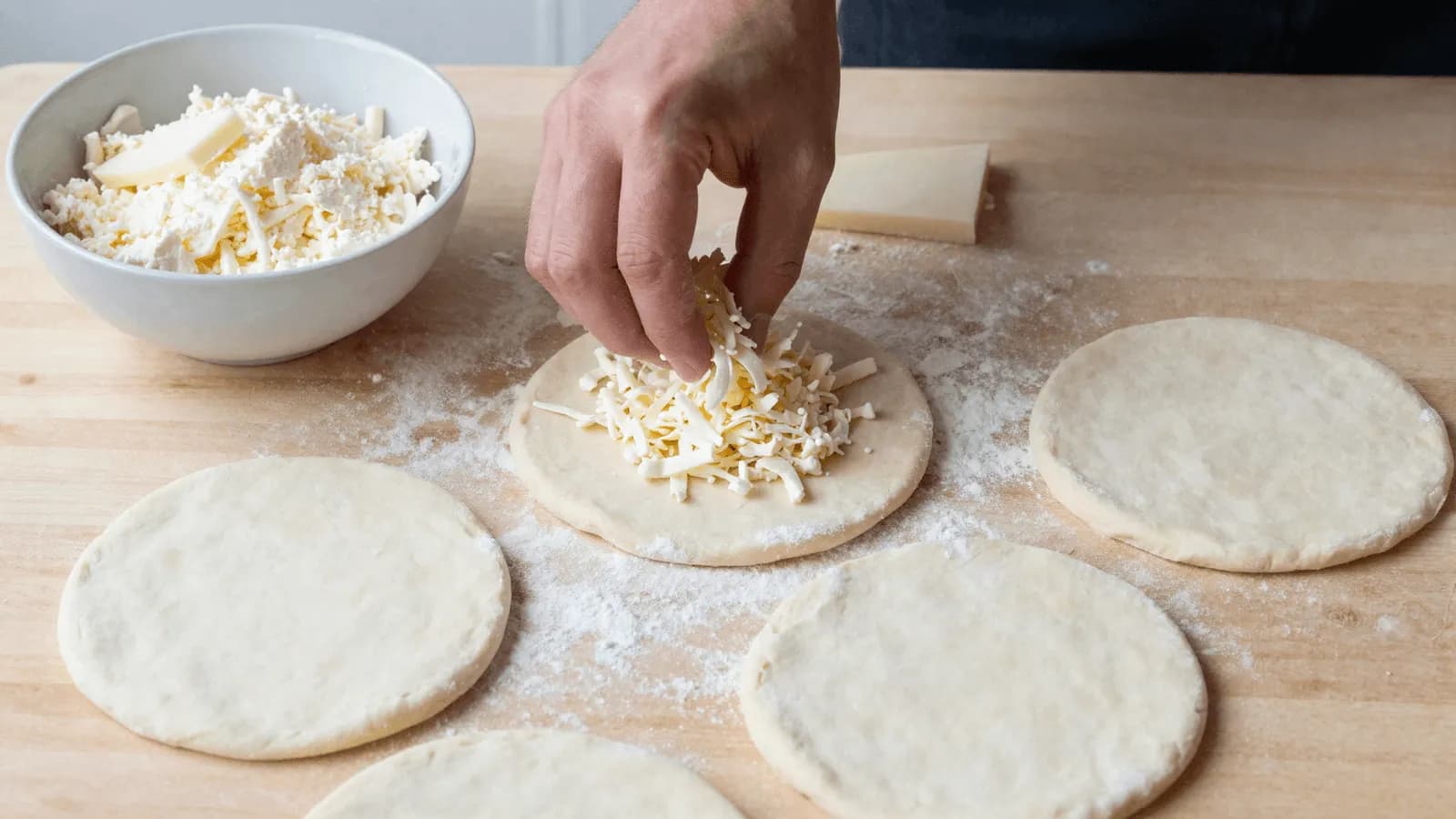 A hand scattering shredded white cheese over raw discs of flatbread dough on a floured wooden board