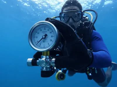 A diver checking a submersible pressure gauge mid-water, light filtering from above