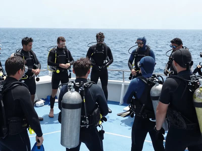 A group of divers gathered around an instructor for a pre-dive briefing on a boat deck