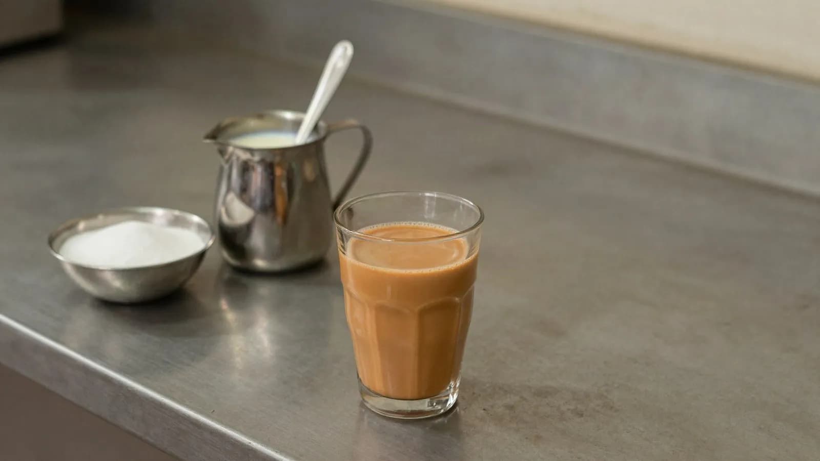 A glass of karak chai beside condensed milk and sugar