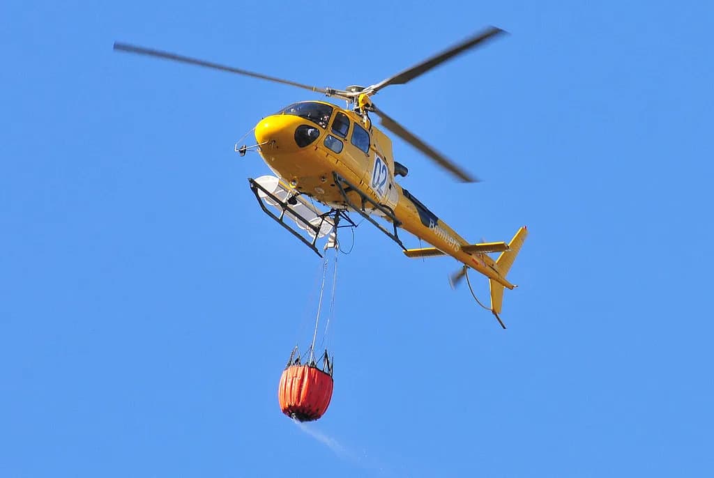 A firefighting helicopter dropping water near a smoke-covered European forest and town