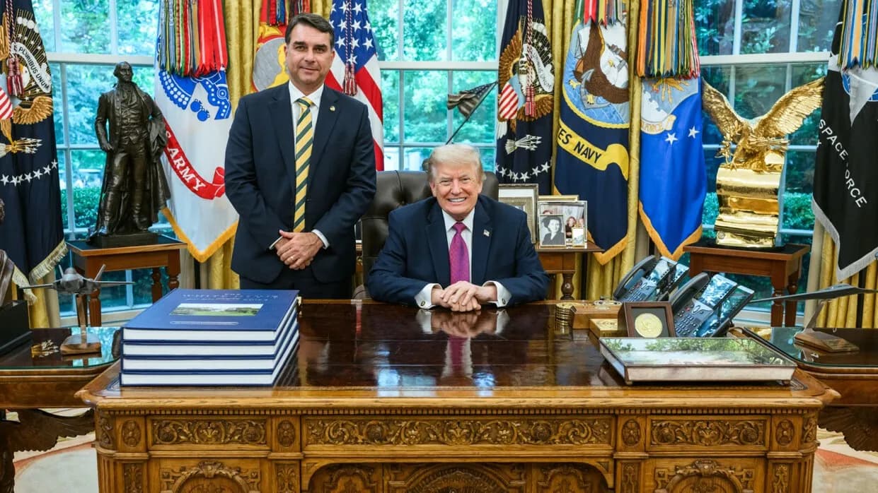 Brazilian Senator Flavio Bolsonaro standing in a dark suit and yellow-striped tie beside a smiling Donald Trump seated at the Resolute Desk in the Oval Office, photographed on 26 May 2026, a file photograph of Bolsonaro and not of his arrival in Miami