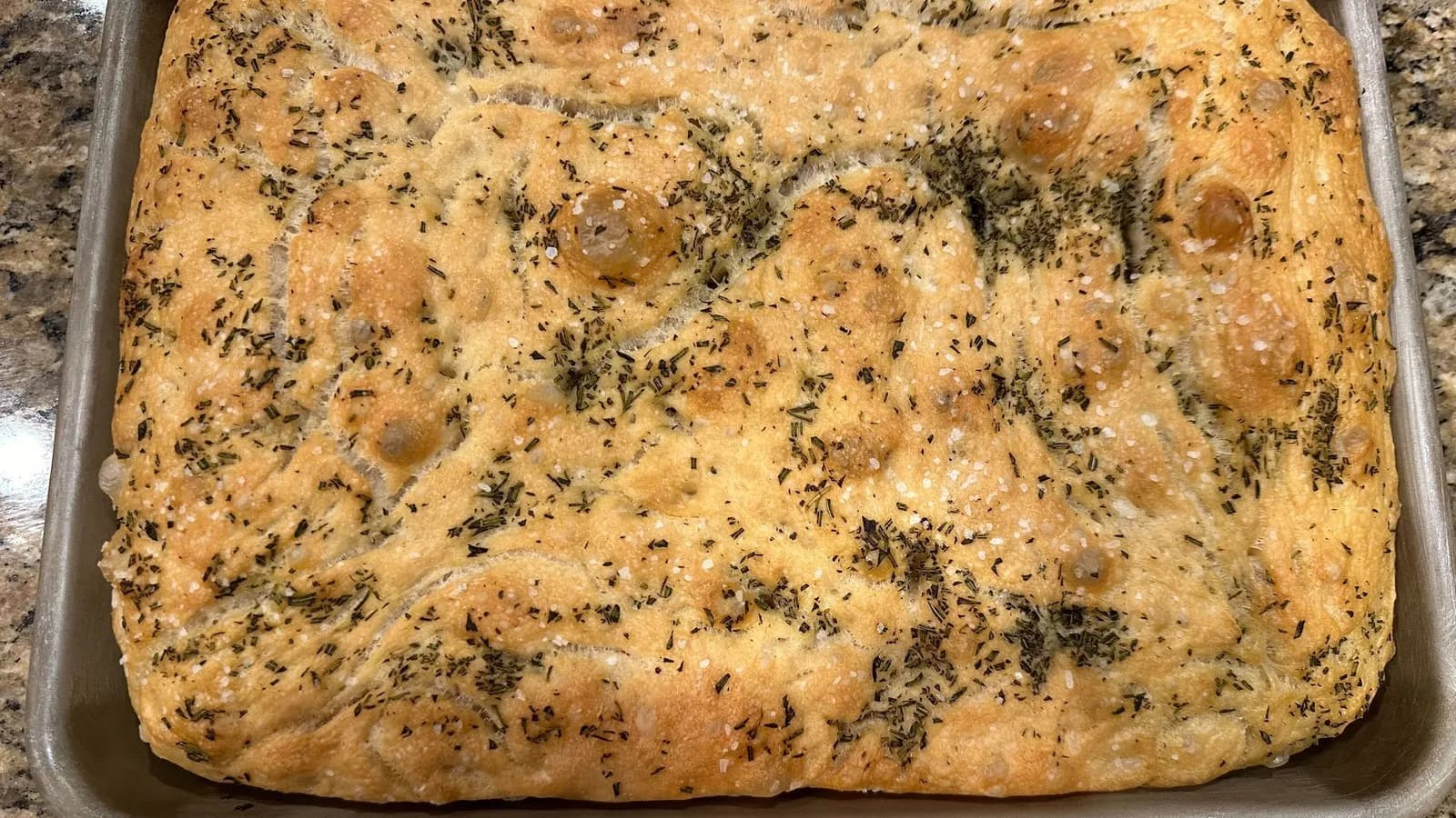 A homemade rosemary focaccia in a metal baking tray, dimpled and golden, baked in the United States and photographed in August 2024
