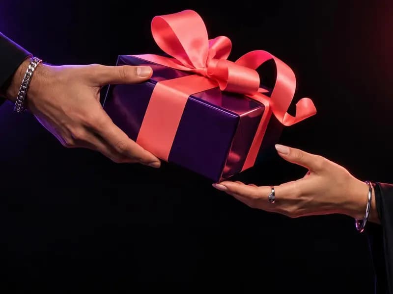 Two hands passing a purple gift box tied with a coral ribbon against a dark background, a Giftze campaign photograph supplied by the company
