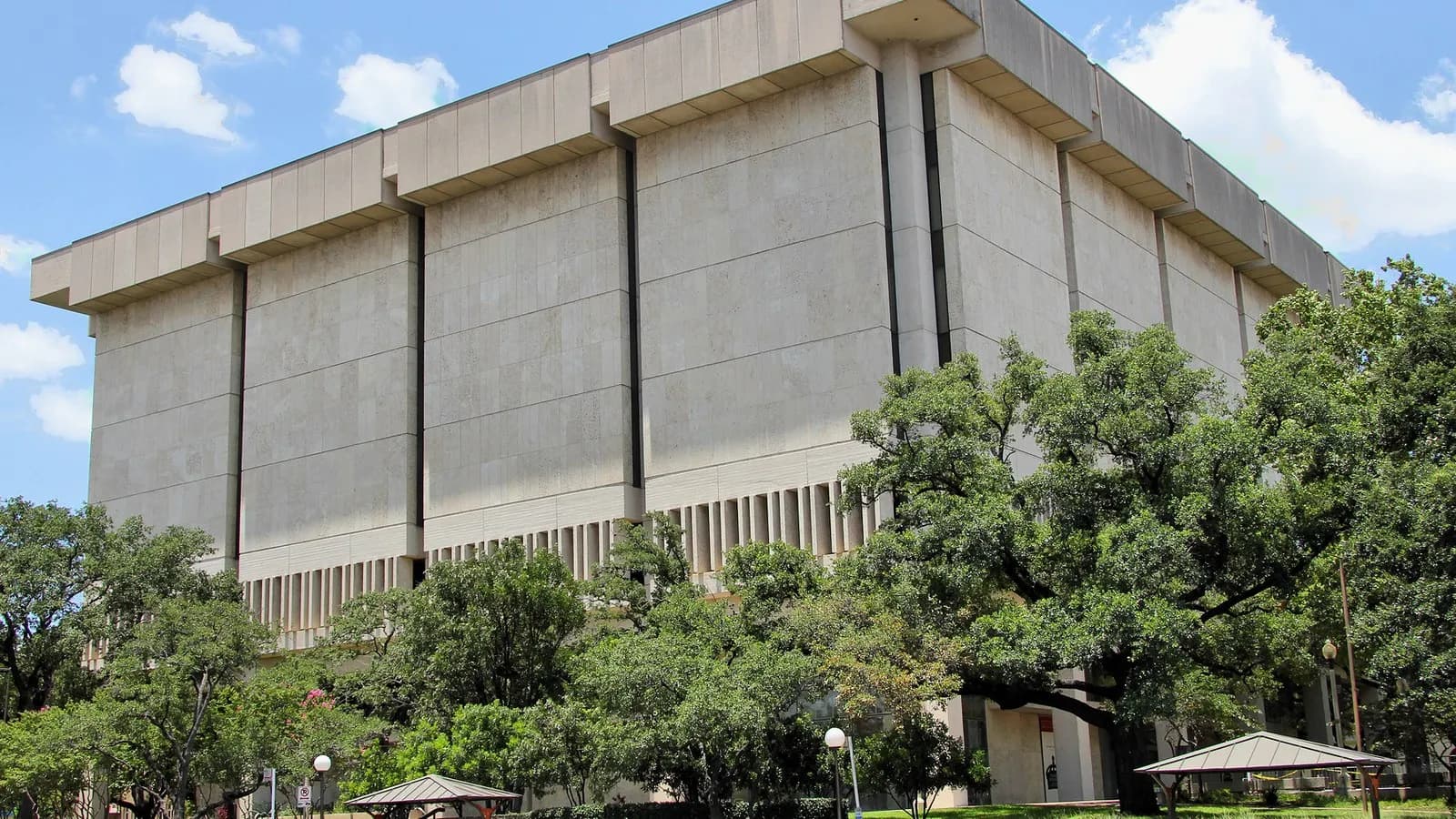 The Harry Ransom Center at the University of Texas at Austin, a tall pale concrete building with long vertical bands, seen from below through green trees under a blue sky, photographed on 16 June 2012, a file photograph of the venue and not of the exhibition