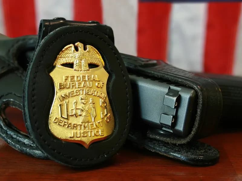 An FBI badge in a leather holder beside a holstered pistol on a wooden desk in front of an American flag, an undated file photograph illustrative of the FBI agents in the series and not a still from it