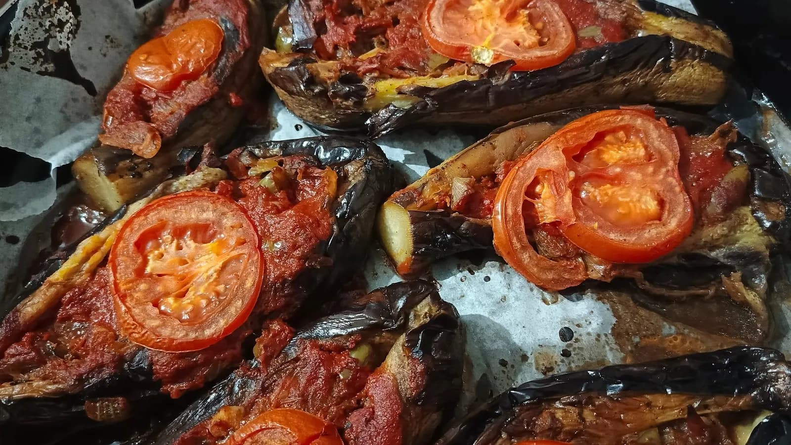 A tray of imam bayildi, whole aubergines split and filled with onion and tomato, each topped with a slice of tomato, glossy from the oil