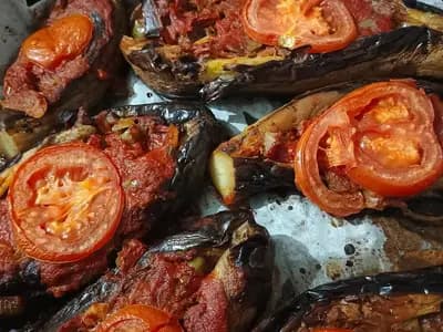 A tray of imam bayildi, whole aubergines split and filled with onion and tomato, each topped with a slice of tomato, glossy from the oil