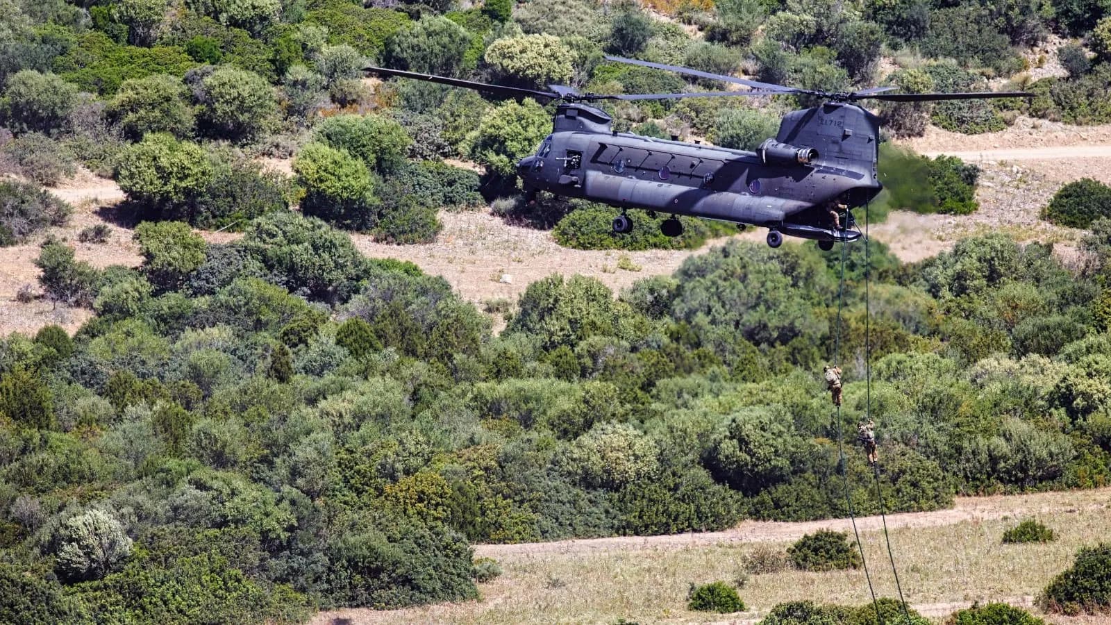 A dark grey Italian Army CH-47F Chinook helicopter of the 1st Army Aviation Regiment Antares hovering over scrubby hillside while personnel descend ropes, photographed on 21 May 2021 during exercise CAEX 1, a file photograph of this helicopter type in Italian service and not of the crash site or of the aircraft involved