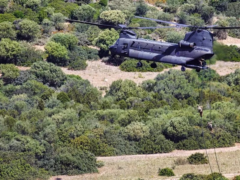 A dark grey Italian Army CH-47F Chinook helicopter of the 1st Army Aviation Regiment Antares hovering over scrubby hillside while personnel descend ropes, photographed on 21 May 2021 during exercise CAEX 1, a file photograph of this helicopter type in Italian service and not of the crash site or of the aircraft involved