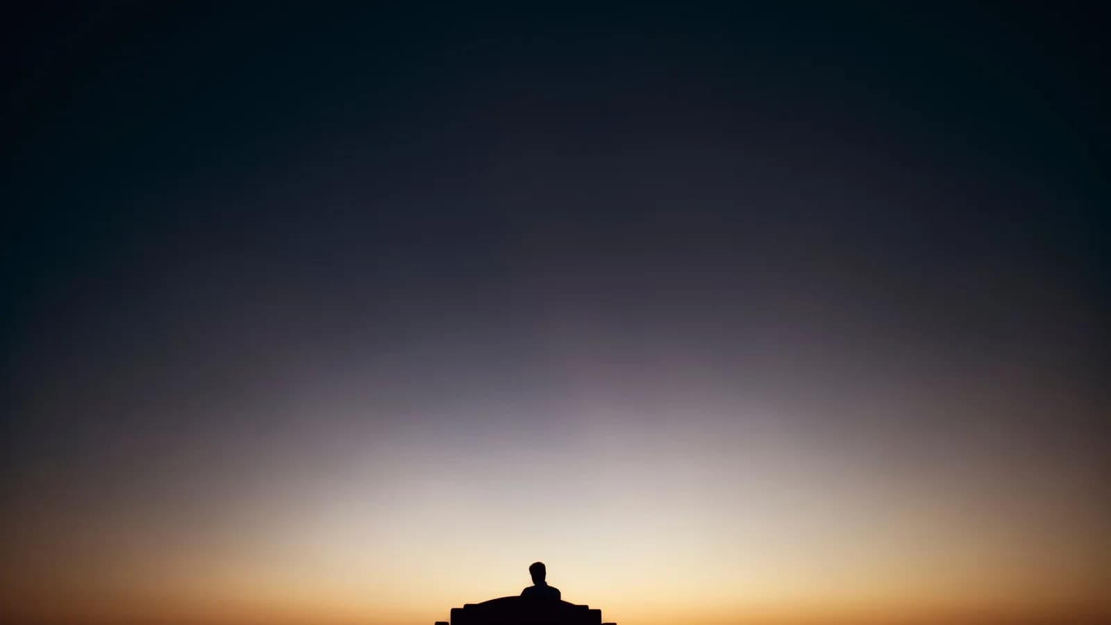 A man sitting alone on a bench in silhouette against a sunset sky, seen from behind