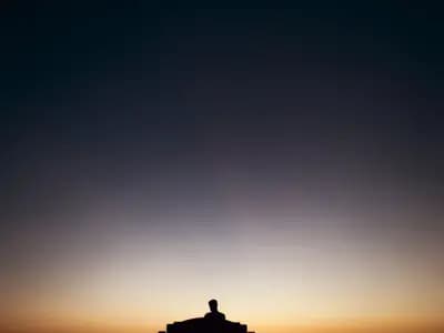 A man sitting alone on a bench in silhouette against a sunset sky, seen from behind