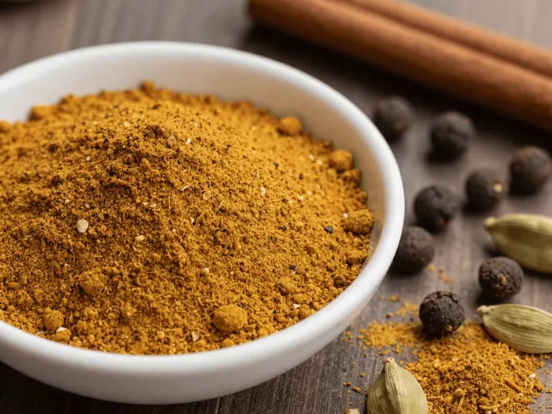 A small bowl of warm brown-gold ground spice blend beside whole allspice berries and a cinnamon stick