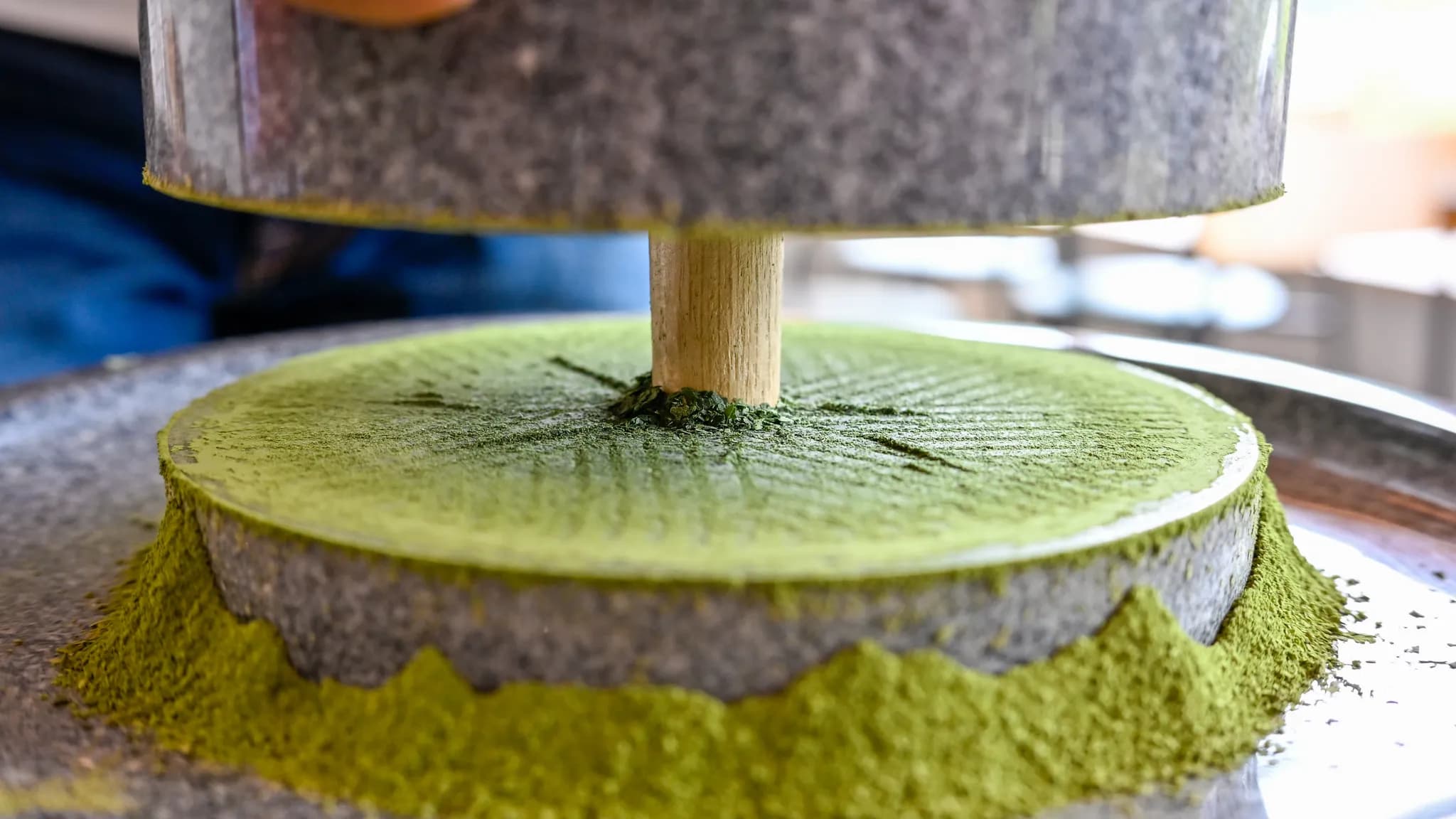 A pale jade green matcha latte being poured into a white ceramic cup on a cafe counter beside a bamboo whisk and tin of matcha powder