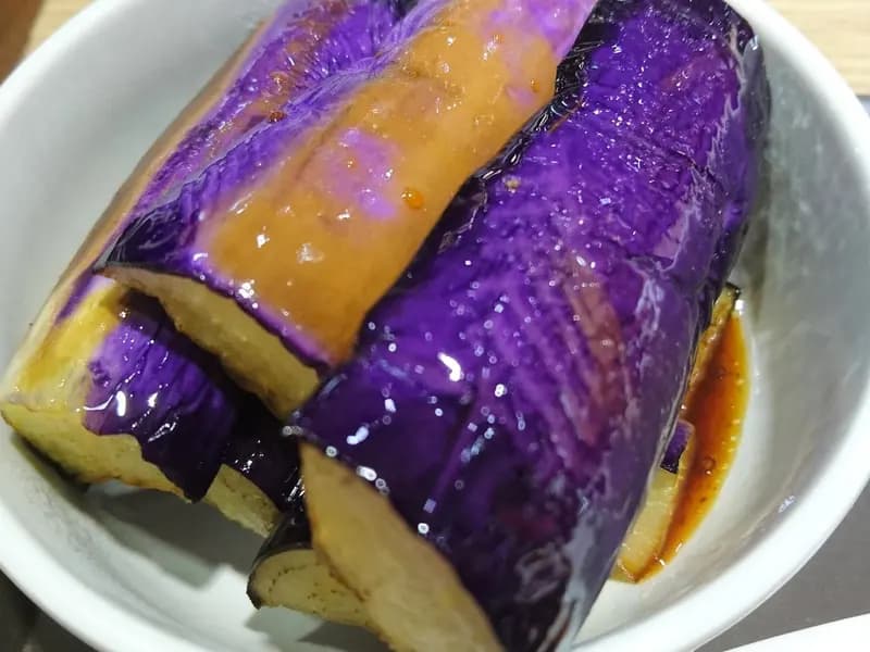 Japanese style grilled aubergine in a shallow bowl, the skin deep purple and the flesh collapsed and glossy under a dark sauce