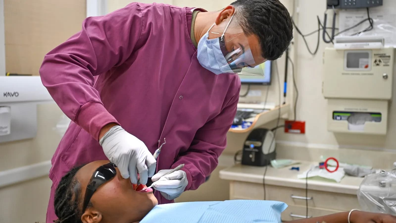 A dental technician in a mask and gloves carrying out an oral examination on a patient lying in a dental chair