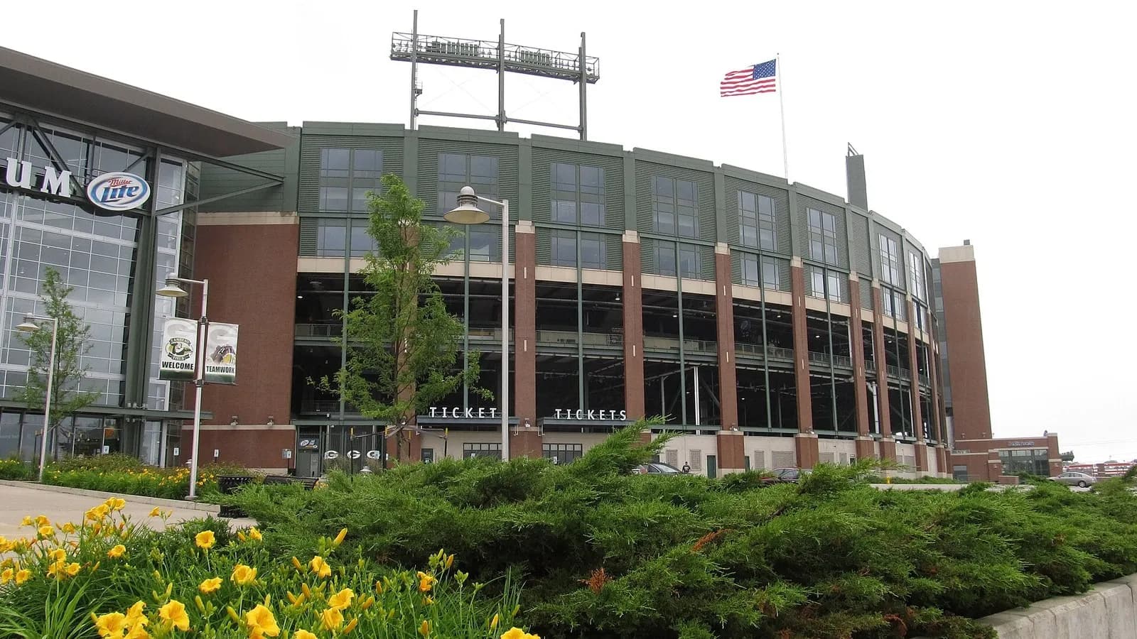 The curved brick and glass exterior of Lambeau Field in Green Bay, Wisconsin, with a flag on the roofline and yellow flowers in the foreground, photographed on 28 June 2013, a file photograph of the Packers' home ground and not of Thursday night's game