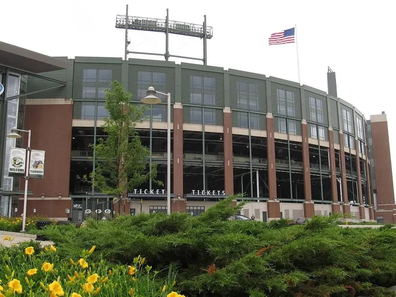 The curved brick and glass exterior of Lambeau Field in Green Bay, Wisconsin, with a flag on the roofline and yellow flowers in the foreground, photographed on 28 June 2013, a file photograph of the Packers' home ground and not of Thursday night's game