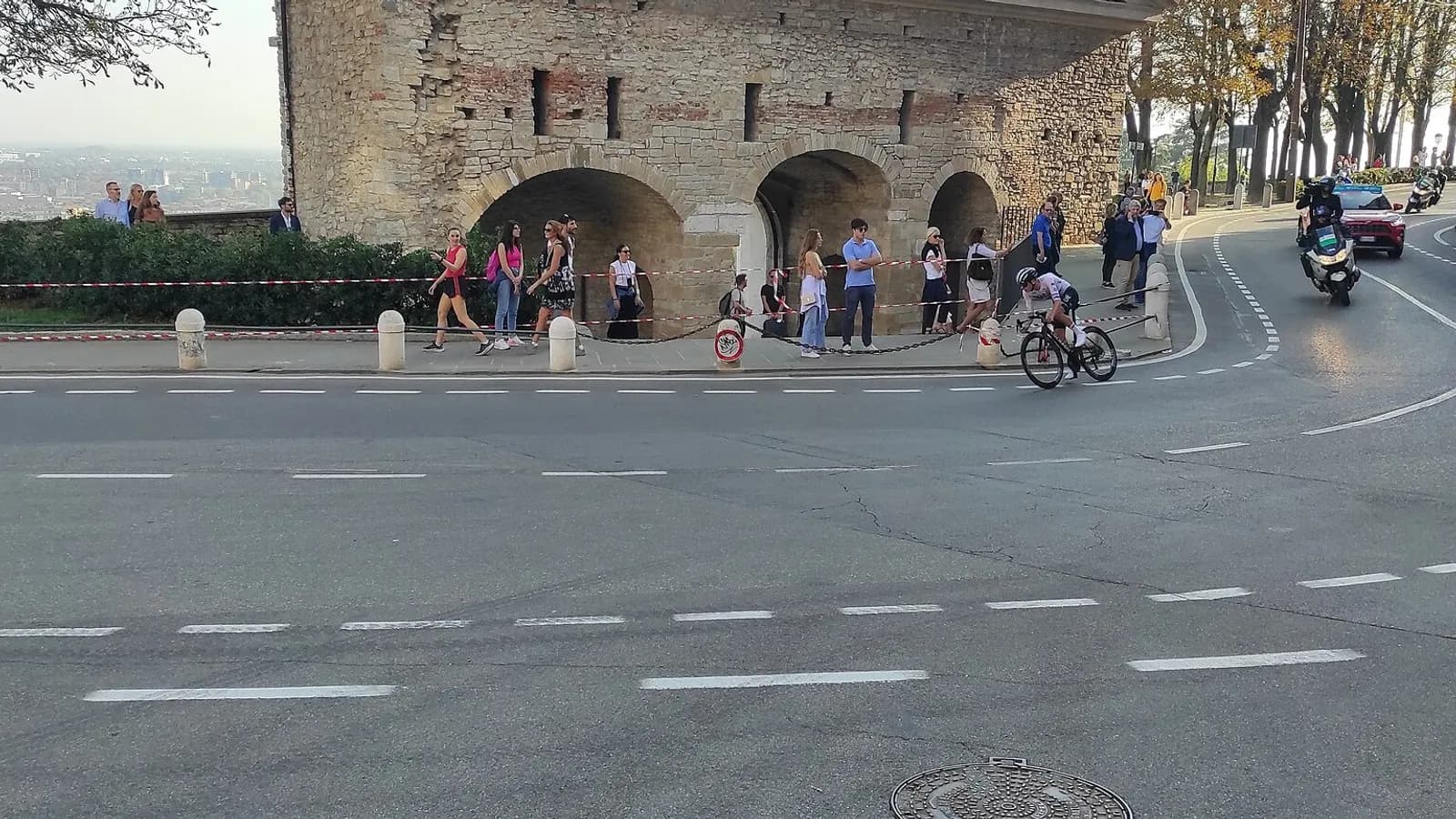 A lone cyclist rounding a bend beneath an old stone wall with spectators and a police motorcycle beside the road, photographed during Il Lombardia on 7 October 2023, a file photograph of the race and not of Paul Seixas or of Saturday's finish